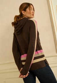 Rudas megztinis su gobtuvu, turintis tekstūruotą paviršių, su rožinėmis, smėlio spalvos ir rudos spalvos juostelėmis apačioje ir rankovėse.