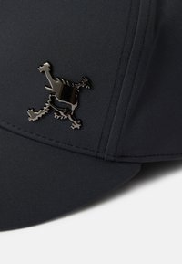 Melnā cepure, izgatavota no gludas auduma, ar mirdzošu melnu logotipu ar sarežģītu detaļām priekšpusē, un izšūtām malām.
