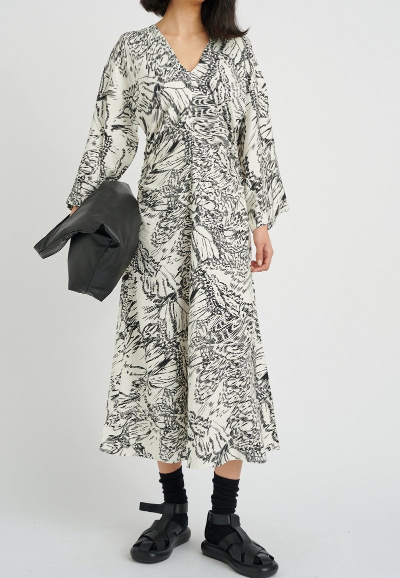 Lång klänning med V-hals i krämfärg med svart abstrakt blommönster. Har vida ärmar och en åtsittande midja. Modellen håller en svart jacka.