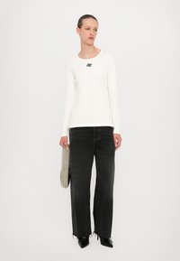 White long-sleeve top with a black logo, paired with high-waisted, wide-leg black jeans. Model holds a beaded clutch and wears black heels.