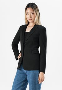 Žena s dlouhými blond vlasy v černém saku a modrých džínech, stojící s jednou rukou na boku na bílém pozadí.