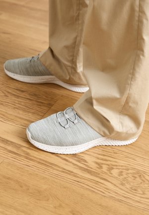 Personne portant des baskets légères gris clair sans lacets et un pantalon beige, debout sur un sol en bois.