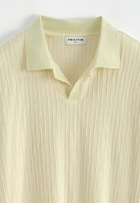 Polo jaune pâle avec un motif texturé, col court et encolure en V, fabriqué dans un tissu doux et lisse. Étiquette taille L visible.