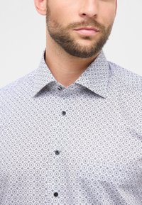 Man met een netjes getrimde baard, gekleed in een wit overhemd met een klein bloemenpatroon, gefotografeerd van de nek tot het midden van de torso op een lichte achtergrond.
