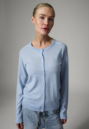 Žena s blond vlasmi nosí svetlomodrý zapínaný sveter a svetlomodré džínsy na jednofarebnom sivom pozadí.