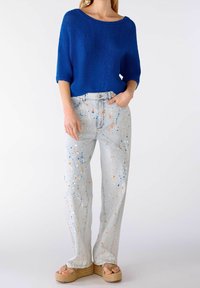 Kobieta ubrana w jasnoniebieski sweter, jasne dżinsy z plamami farby w kolorze pomarańczowym, niebieskim i białym oraz sandały na platformie z korkowymi podeszwami.