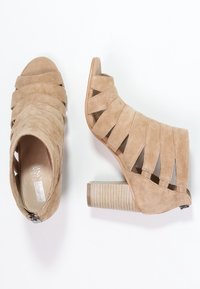 Beige nubuck avokkaat, joissa on avonaiset varpaat, ristikkäiset hihnat, teksturoitu korko ja sivuvetoketju. Littee, uritettu kenkäkorko.