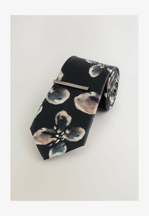 Modeļots zīda kaklasiksna ar tumši zilu fonu un abstraktiem gaišiem ziedu rakstiem. Iekļauta sudraba kaklasiksnas skava drošai nostiprināšanai.