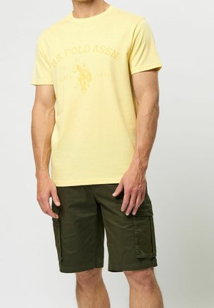 Mann trägt ein blassgelbes U.S. Polo Assn. T-Shirt und dunkelgrüne Cargoshorts, steht vor einem einfarbig hellen Hintergrund.