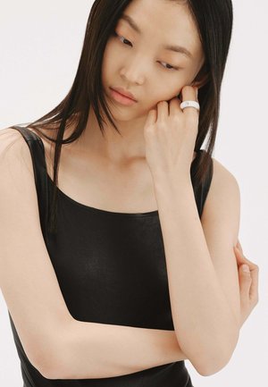 Top negro sin mangas con una textura suave, combinado con un anillo blanco que presenta un diseño sutil. La mano del modelo reposa sobre su mentón.