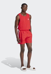 Κόκκινο μπλουζάκι με λευκό λογότυπο, συνδυασμένο με αντίστοιχες κόκκινες βερμούδες που διαθέτουν τρεις λευκές ρίγες στα πλάγια. Φορώντας λευκά σαγιονάρες.