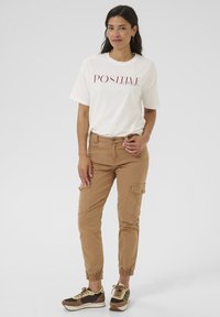 Bílý tričko s červeným nápisem "POSITIVE", khaki cargo kalhoty a hnědé tenisky se světlými akcenty. Casual střih, ukazující kapsy a elastické manžety.