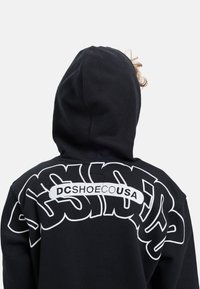 Čierna mikina s kapucňou s veľkým bielym grafickým motívom na chrbte, ktorý zobrazuje nápis "DCSHOEUSA". Dizajn zahŕňa výrazné, štýlové písanie a kapucňu.