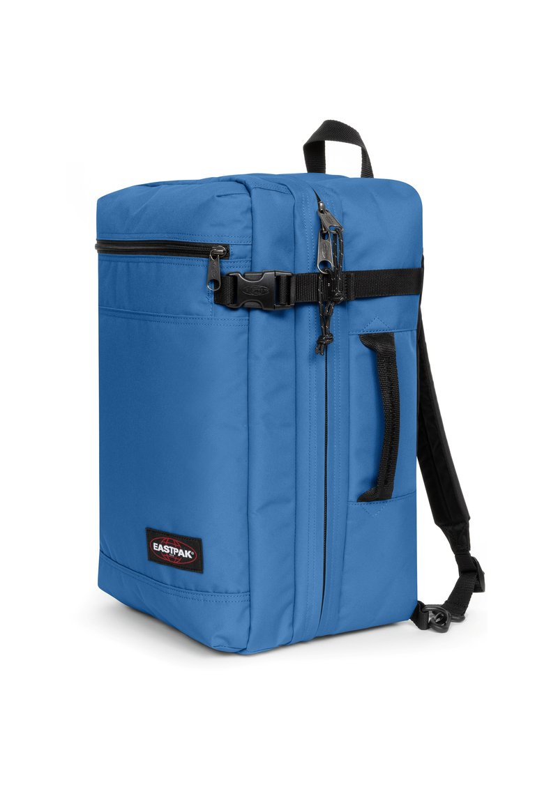 Mochila rectangular azul con una textura de tela suave, cremalleras, bolsillo lateral y correas ajustables negras. Parche con el logo en la base.