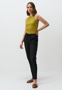 Olijfgroen geribbeld mouwloos topje gecombineerd met zwarte broek met trekkoord. Model draagt bruine sliders. Minimalistisch ontwerp, gladde textuur.