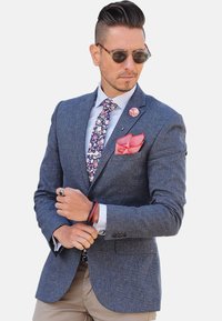 Mørkeblå struktureret blazer, hvid skjorte, blomstercravat, rød lommeklud og solbriller. Bemærkelsesværdige detaljer inkluderer en reversnål og armbånd.