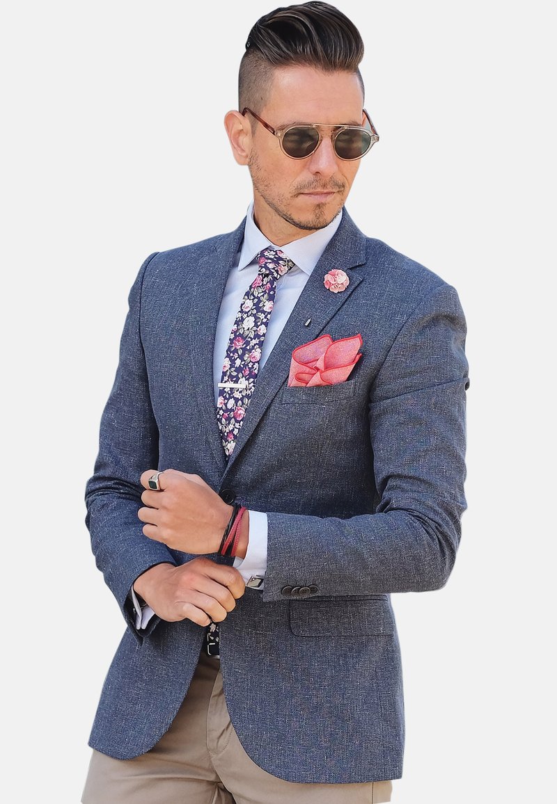 Mørkeblå struktureret blazer, hvid skjorte, blomstercravat, rød lommeklud og solbriller. Bemærkelsesværdige detaljer inkluderer en reversnål og armbånd.