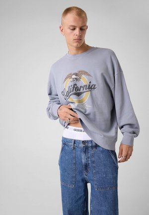 Jeune homme portant un sweat gris California, un jean bleu et une ceinture blanche Guess, soulève le sweat pour révéler le nom de la marque.