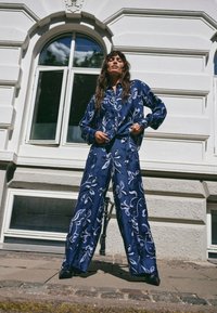 Tmavě modrá saténová košile s květinovým vzorem a široké kalhoty s dlouhými rukávy a volným střihem, nošené s černými lodičkami.