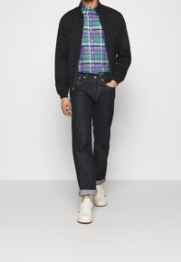 Black bomber jacket, plaid button-up shirt in green, purple, and blue, dark denim jeans, and white sneakers with light brown soles.