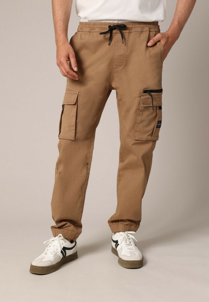 Pantalon cargo beige avec taille élastique et cordon de serrage ajustable. Comprend plusieurs poches, dont une poche latérale zippée.
