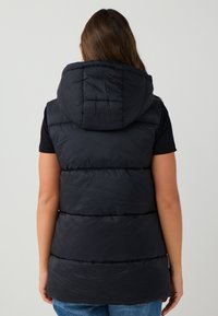 Gilet puffer senza maniche di colore nero con cappuccio, presenta dettagli trapuntati e una texture liscia, abbinato a una semplice t-shirt nera sottostante.