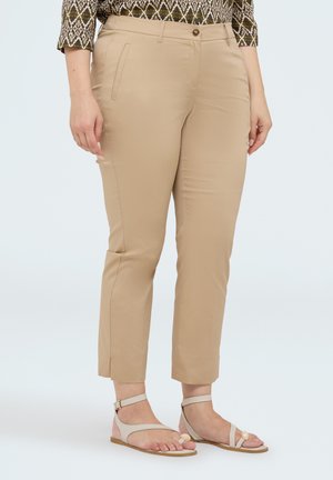Vrouw draagt beige slim-fit enkelbroeken en witte platte sandalen met enkelbandjes, staand tegen een effen lichte achtergrond.