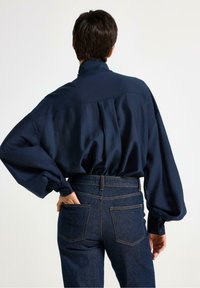 Marineblå oversized bluse med bølgende ærmer, knapmanchetter og høj krave, parret med mørkeblå jeans med baglommer.
