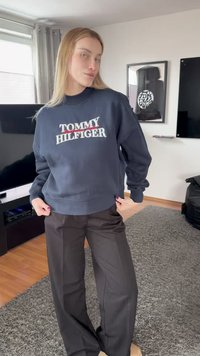 Νεαρό άτομο με μακριά ξανθά μαλλιά φοράει μπλε σκούρο φούτερ Tommy Hilfiger και μαύρο φαρδύ παντελόνι στέκεται σε σαλόνι με τηλεόραση και χαλί.