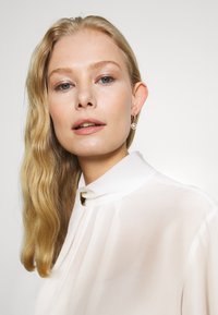 Rambut pirang dengan gelombang lembut, mengenakan blus putih berkerah tinggi. Memiliki riasan yang lembut dan sepasang anting jatuh yang berkilau.
