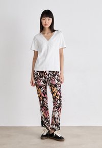 Weißes kurzärmliges Hemd mit gesticktem Schmetterlingsdesign, kombiniert mit farbenfrohen gemusterten Hosen in Pink-, Orange- und Schwarz-Tönen. Schwarze Schuhe.