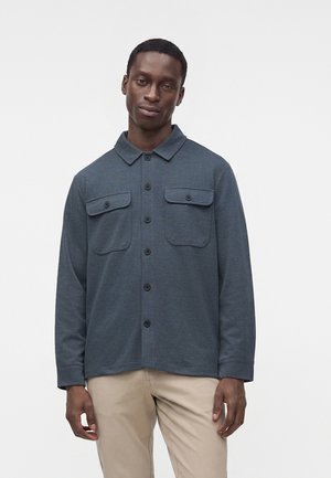 Mand iført en mørkegrå skjorte med knapper og to forlommer og beige bukser, der står mod en ensfarvet lys baggrund.