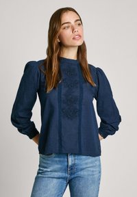 Blusa blu navy con maniche a sbuffo lunghe, ricamo al centro e pizzo attorno al collo e ai polsini. Tessuto in cotone.