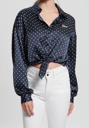 Woman wearing a navy blue satin polka dot long-sleeve shirt tied at the waist, paired with high-waisted white jeans.