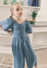 Lysblå denim jumpsuit med puffærmer og snøre i taljen. Har samlet detaljer på brystet og vide bukser.