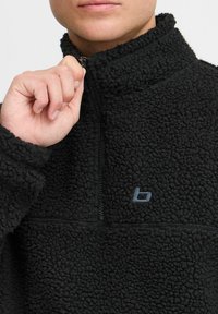 Pull en polaire noir avec un col haut, fermeture éclair quart et tissu texturé. Présente un petit logo gris sur la poitrine.