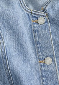 Svijetloplava denimska tkanina prikazuje izbliza dizajn s ovratnikom, metalnim kopčama i kontrastnim žutim šavovima.