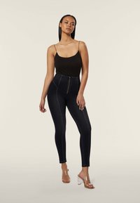 Leggings in denim scuro a vita alta con zip frontale, cuciture a contrasto e silhouette aderente, abbinati a una canotta nera e tacchi trasparenti.