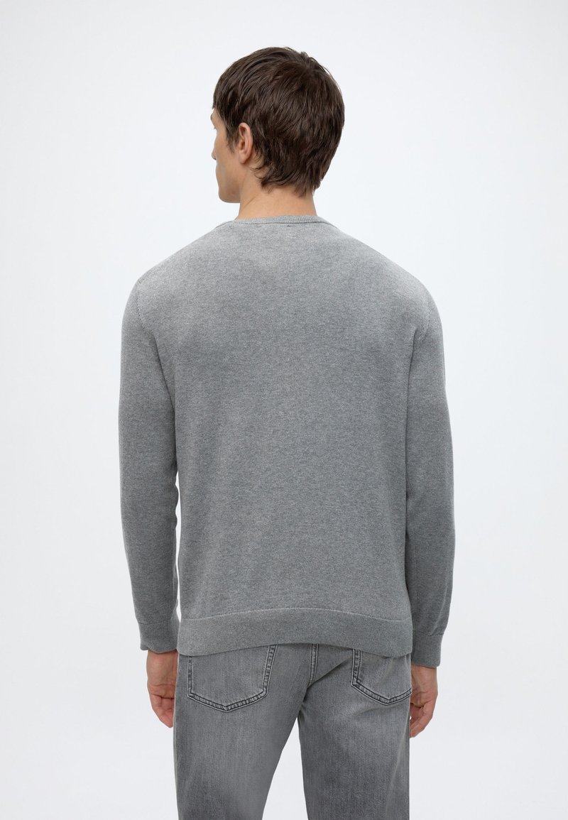 Man wearing a plain grey long-sleeve sweater and grey jeans, standing with his back facing the camera on a white background.