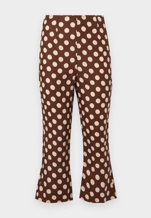 Frau mit langen, glatten schwarzen Haaren, die ein trägerloses braunes Top und kurze Hosen mit großen weißen Punkten trägt, steht mit einer Hand in der Hüfte.