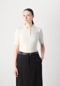 Kortsleeve'i ribblikuga elevandiluust värvitud polo särk, millel on kolm nööpi, koos kõrge vöökohaga mustade pükste ja hõbedase pandlaga musta vöödiga.