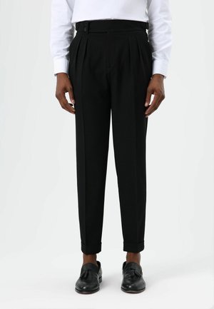 Pantalon noir plissé à taille haute, avec des fermetures à boutons et une coupe ajustée. Porté avec des mocassins noirs et une chemise blanche.