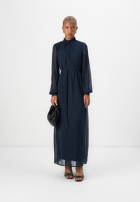 Μακρύ φόρεμα ναυτικού μπλε με μακρές διαφανείς μανίκες, ψηλή λαιμόκοψη, ζώνη στη μέση και ρέουσα ύφασμα, συνδυασμένο με μαύρη τσάντα και στολισμένα σανδάλια.