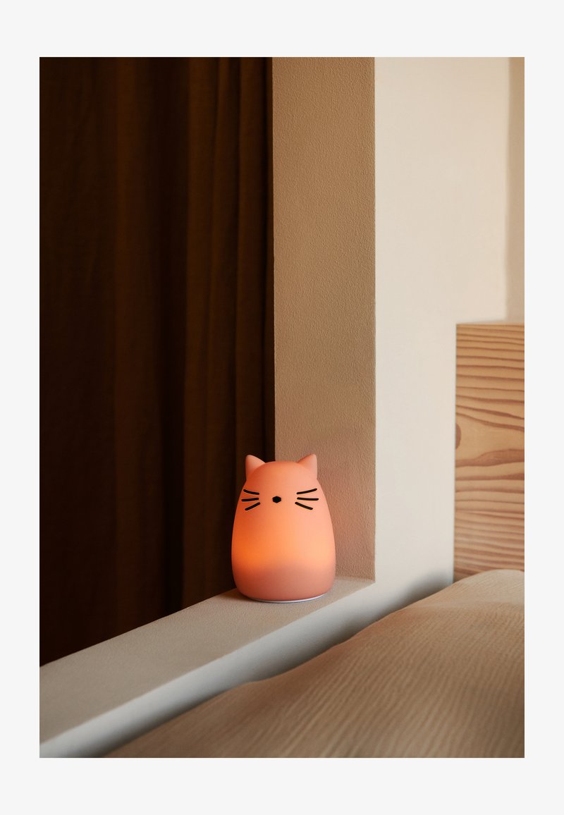 Kattformad nattlampa i mjuk rosa silikon, med svarta morrhår och en minimalistisk design, placerad på en vit hylla bredvid en säng av trä.