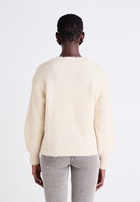 Cream-colored knitted sweater with a ribbed texture, drop shoulders, and a relaxed fit, paired with gray striped pants.