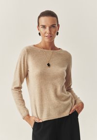 Beige kasjmier trui met een ronde hals, geribbelde zoom en lange mouwen. Gecombineerd met een zwarte hangende ketting en ronde zwarte oorbellen.