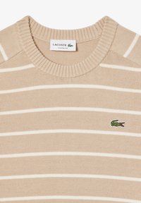 Beige pletený svetr s bílými vodorovnými pruhy, s žebrovaným kulatým výstřihem, na levé straně s malým zeleným logem Lacoste. Klasický střih.
