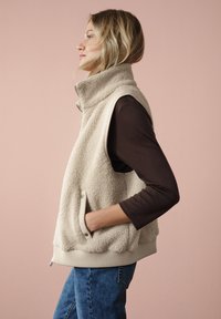 Beige fleece vest met hoge kraag, zijzakken en geribde rand. G gedragen over een bruin langemouwen top, gecombineerd met blauwe jeans.