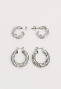 Divas divas sudraba auskari: viens pāris ar teksturētu lapas dizainu, otrs ar apļveida formu, rotāts ar spīdošiem dimantiem.