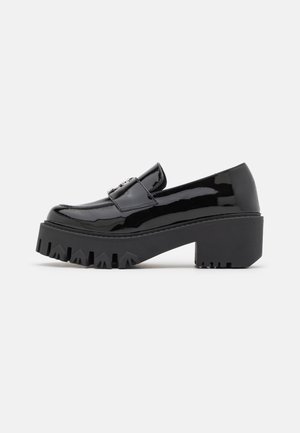 Mocassin en cuir verni noir avec semelle crantée épaisse et talon bloc, orné d'une boucle métallique décorative sur le dessus.